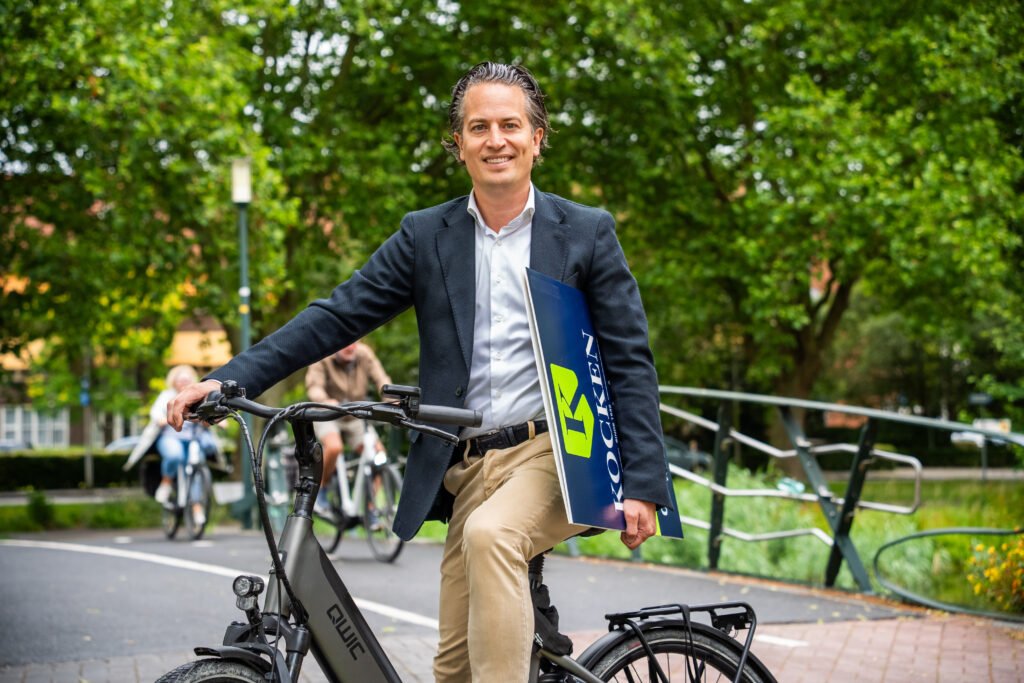 Makelaar van Kocken Makelaars op de fiets met een te koop bord onder de arm in een groene straat in Purmerend