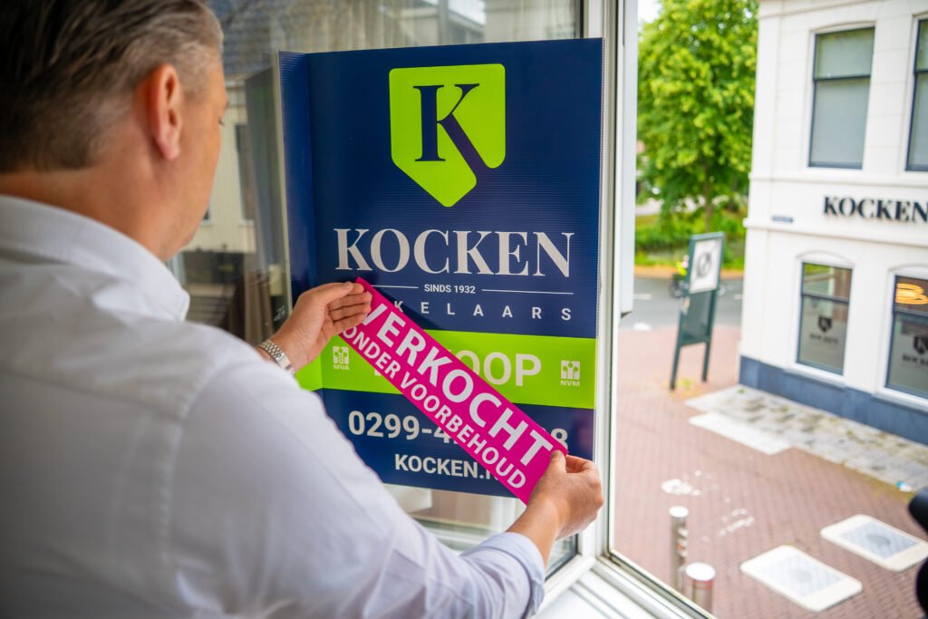 Makelaar plakt een sticker Verkocht onder voorbehoud op een te koop bord van Kocken Makelaars in Purmerend