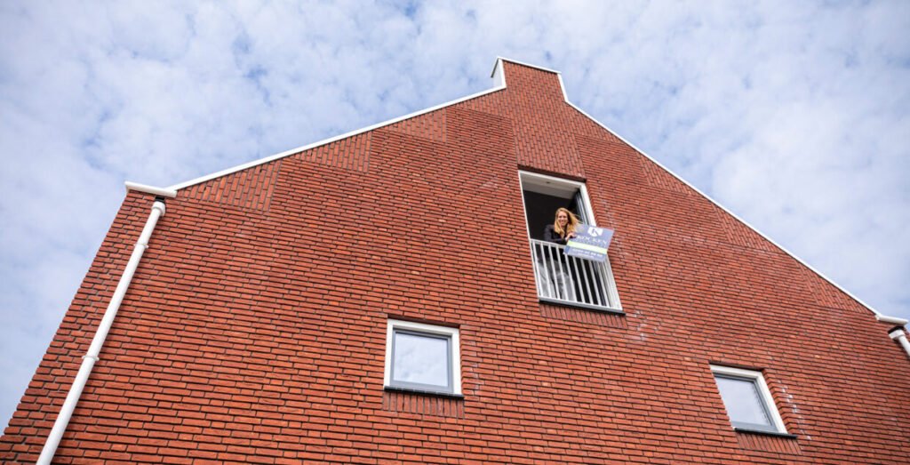 Vrouw leunt uit het raam van een bakstenen woning met een bord van Kocken Makelaars, voor de zoekopdracht naar een nieuw huis
